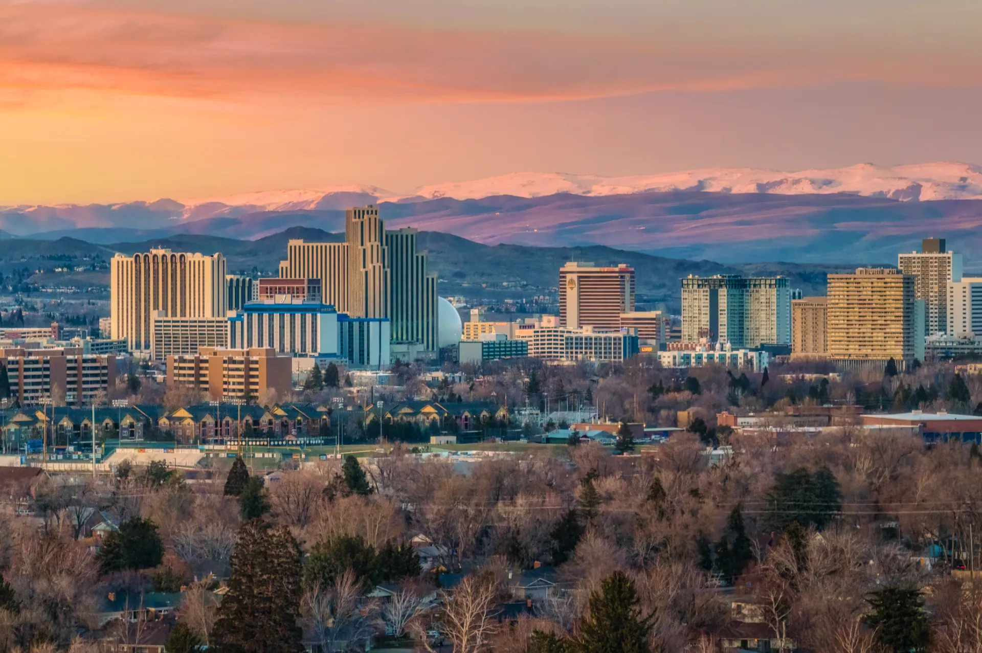 Downtown Reno's casino towers at sunrise, with snow on the Sierra Nevada behind them and tree-lined residential streets in the foreground